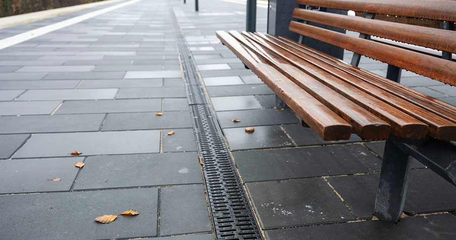 Leerer Bahnsteig mit Pflastersteinen, Entwässerungsrinne, Holzbank im Vordergrund und herbstlichen Blättern auf dem Boden.