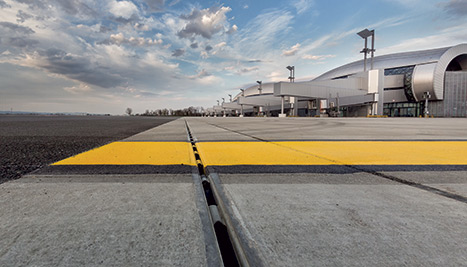 Flughafenvorfeld mit gelbem Markierungsband und Entwässerungsrinne in der Betonfläche, Terminal im Hintergrund.