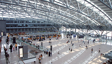 Große, helle Flughafenhalle mit vielen Reisenden, Check‑in‑Schaltern und sichtbarer Stahlfachwerk‑Decke.