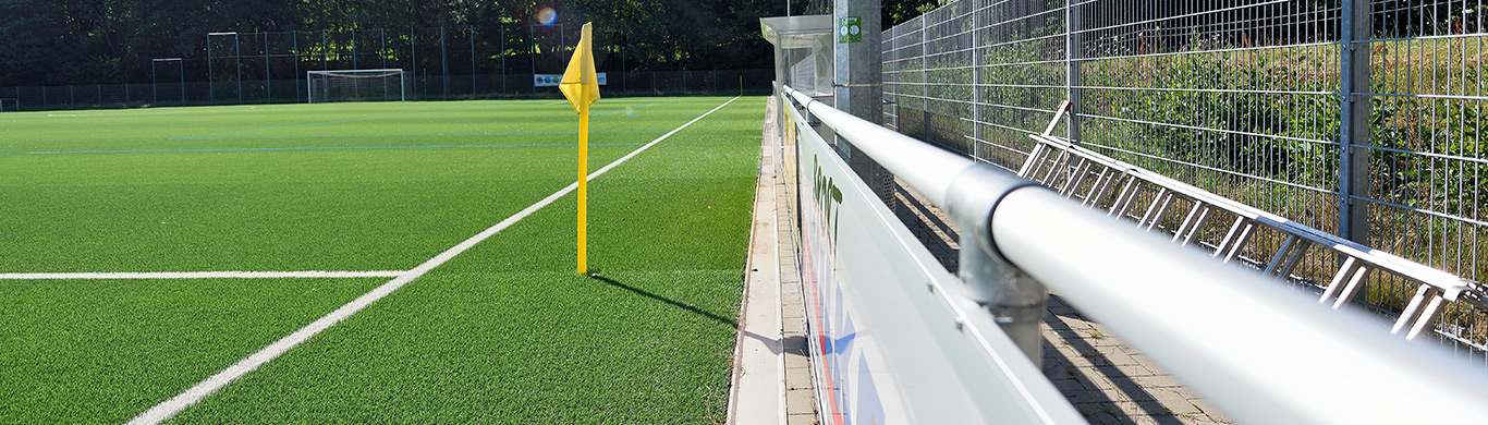 Sportplatz mit grünem Kunstrasen, weißen Spielfeldmarkierungen und roter Laufbahn, umgeben von Bäumen und Tribünen.