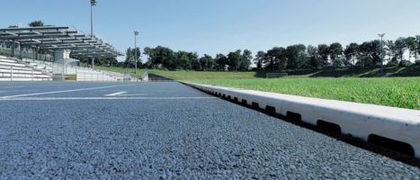 Blaue Laufbahn neben Sportplatz mit Rasenfläche. Im Hintergrund eine Tribüne.