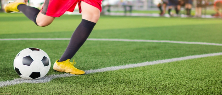 Mann mit roter Sporthose und gelben Fußballschuhen tritt Fußball auf Rasenplatz.