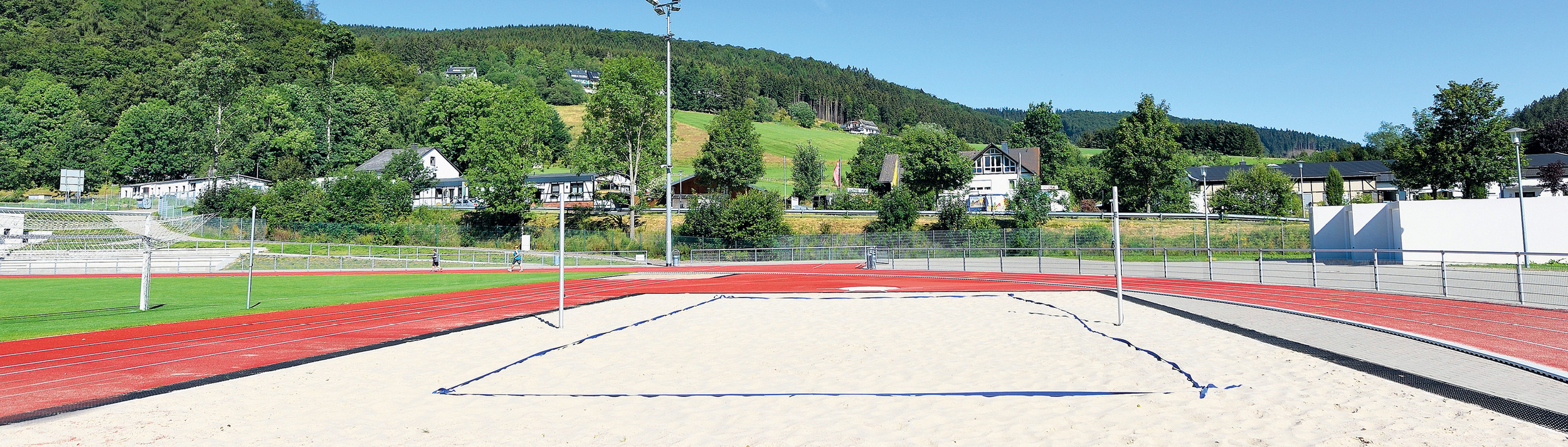 Volleyballplatz draußen umgeben von roter Tartanbahn und Rasenfläche. Im Hintergrund hügelige Wälder und Häuser.