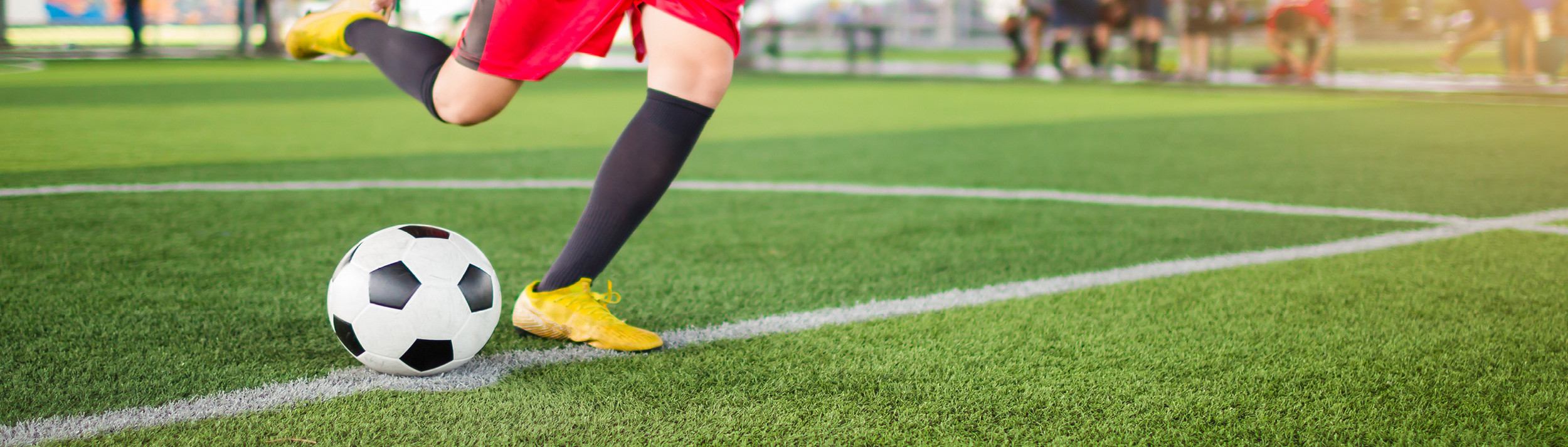 Mann mit roter Sporthose und gelben Fußballschuhen tritt Fußball auf Rasenplatz.