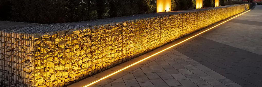 Beleuchtete Steinmauer mit warmem Licht entlang eines gepflasterten Gehwegs bei Dämmerung, mit Laternen und Büschen im Hintergrund.