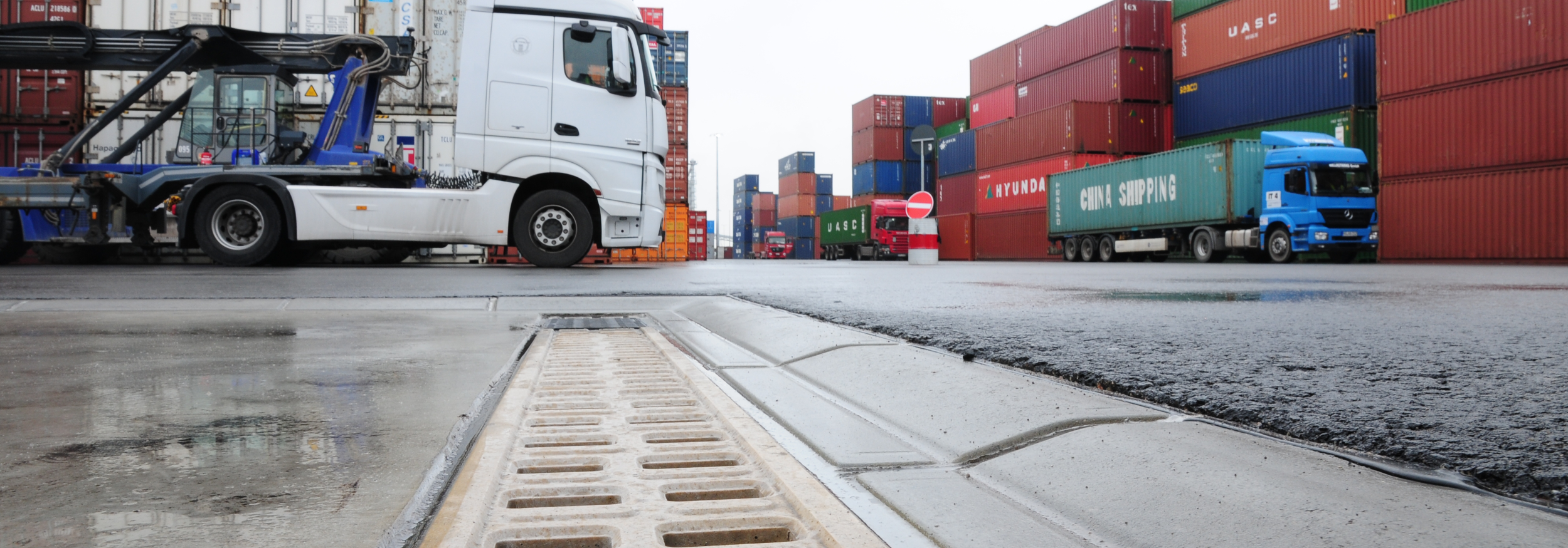 Entwässerungssysteme im Schwerlastbereich LKWs und Container stehen auf einem Hafenplatz mit Entwässerungsrinne im Vordergrund.