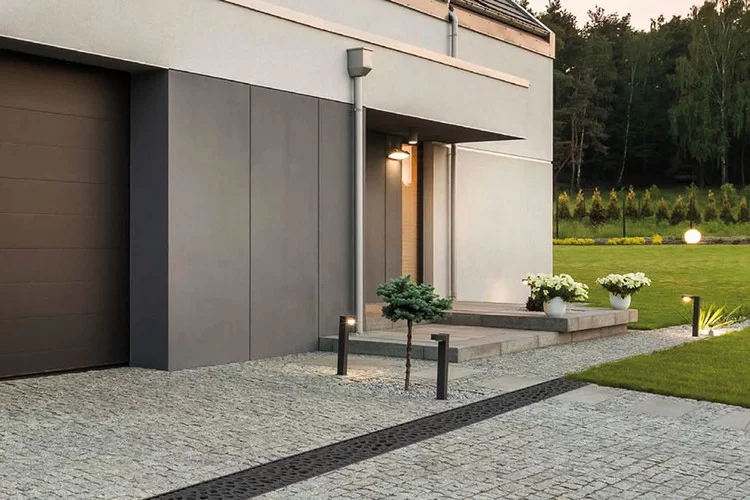 Moderner Hauseingang mit heller Fassade, gepflasterter Einfahrt mit Linienentwässerungsrinne, Gartenleuchten und Bepflanzung.