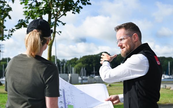Zwei Personen betrachten einen großen Bauplan im Freien, mit Bäumen und einem Hafen im Hintergrund.