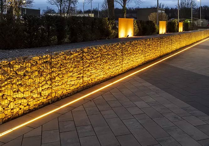Beleuchtete Linienentwässerungsrinne mit LED-Technik entlang einer Gabionenwand bei Dämmerung, Büsche im Hintergrund.