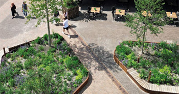 Vogelperspektive auf einen gepflasterten Platz mit zwei bepflanzten Inseln, Bänken und wenigen vorbeigehenden Personen.