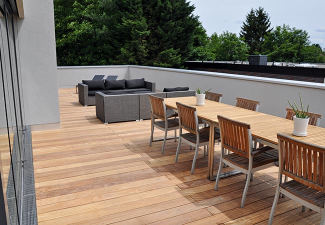 Fassadenrinnen vor einer Schiebetür auf Holzterrasse, langer Esstisch mit Stühlen und Lounge-Bereich.