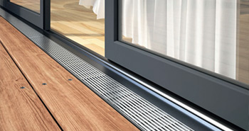 Bodennahes Foto einer Fassadenrinnen mit Schlitzrinnen vor einer leicht geöffneten Schiebetür auf Holzterrasse.