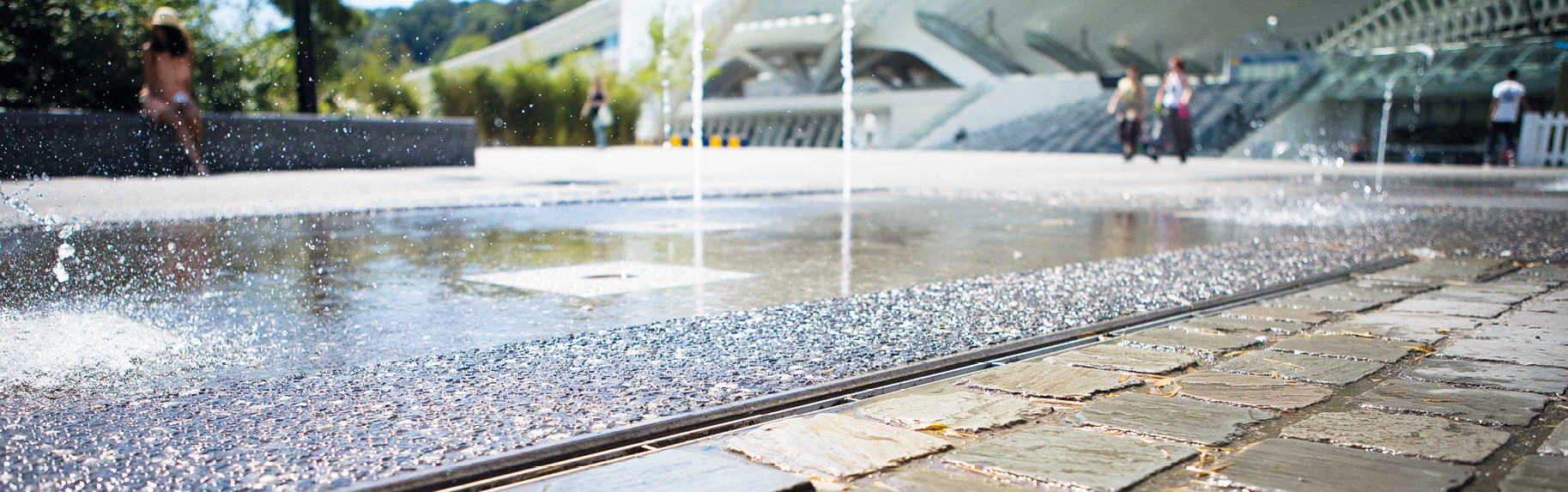Moderner Stadtplatz mit flachem Springbrunnen, sprudelndem Wasserstrahl und umliegenden Sitzbereichen.
