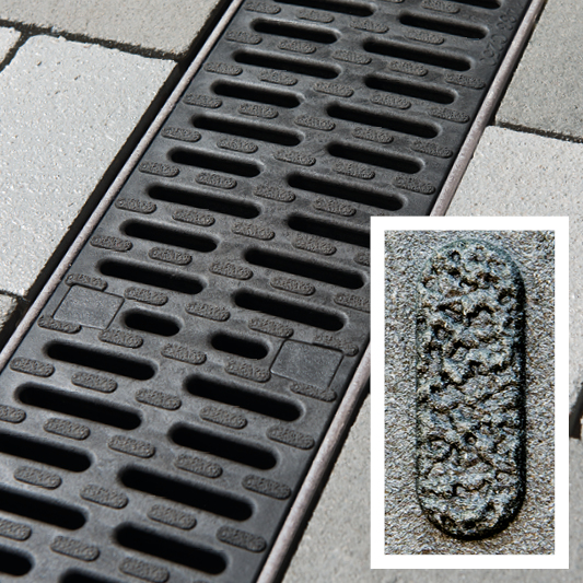 Nahaufnahme einer schwarzen Entwässerungsrinne mit Rost in gepflasterter Fläche, Detailansicht des Oberflächenmaterials.