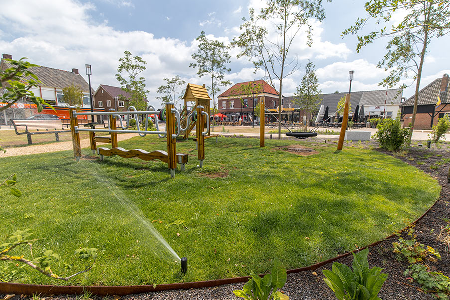 Grüne Spielfläche mit Holzspielgeräten und Sprinklerbewässerung in einem modernen Park, umgeben von jungen Bäumen und Häusern.