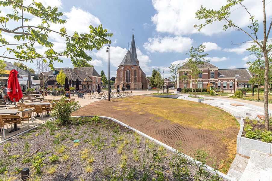 Moderner, sonniger Stadtplatz mit Sitzbereichen, Außengastronomie, jungen Pflanzen und Blick auf eine Kirche im Hintergrund.