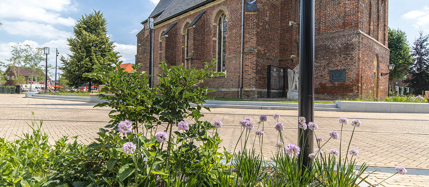 Innerstädtischer Platz mit Pflanzenbeet vor einer historischen Backsteinkirche bei sonnigem Wetter.