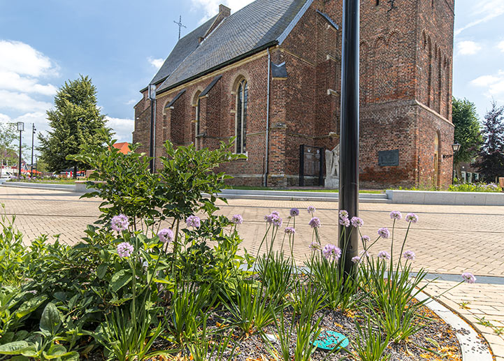Innerstädtischer Platz mit Pflanzenbeet vor einer historischen Backsteinkirche bei sonnigem Wetter.