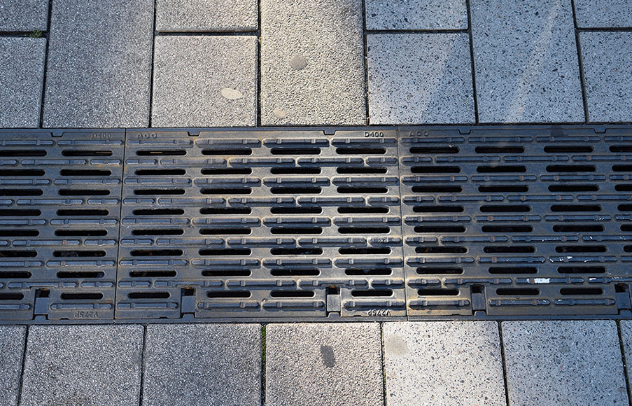 Detailansicht einer Entwässerungsrinne mit taktiler Leitstruktur in gepflasterter Fläche.
