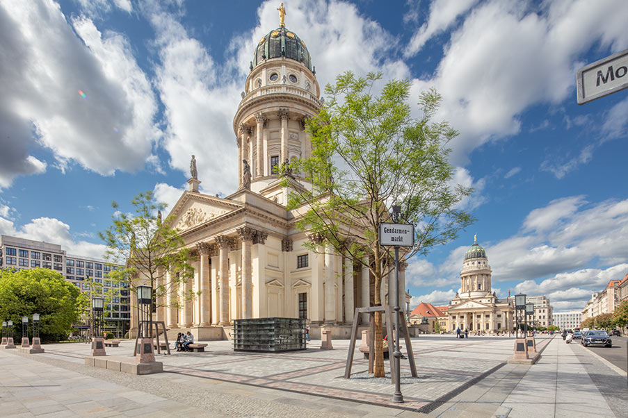 Der Deutsche Dom auf dem Gendarmenmarkt in Berlin, mit einer großen Platzfläche unter einem blauen Himmel mit Wolken.