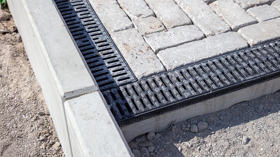 Schwarze Entwässerungsrinne mit Rost in einer gepflasterten Fläche, eingefasst von Betonrand.