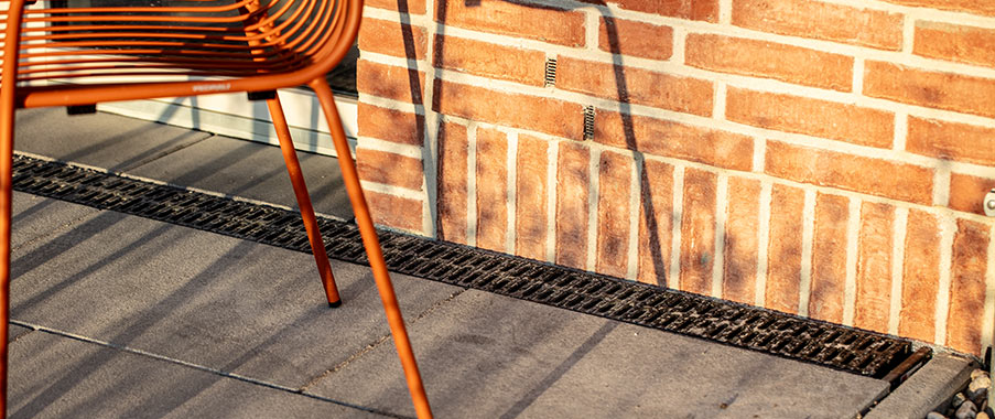 Terrassenbereich mit grauen Platten, orangefarbenem Stuhl und schwarzer Entwässerungsrinne entlang einer Backsteinwand.