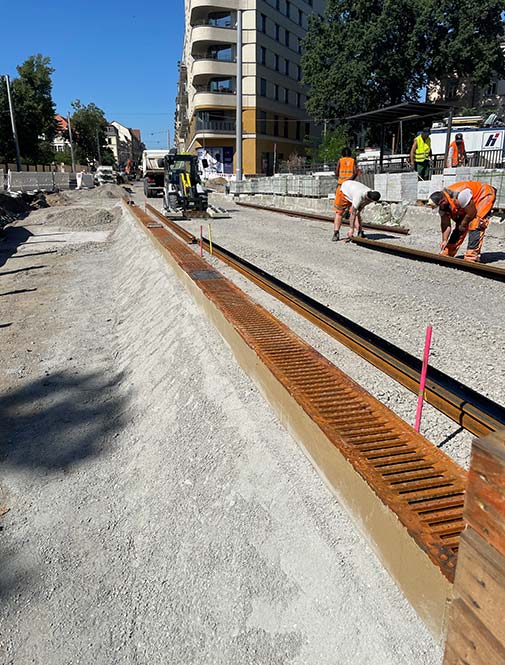 Bauarbeiten auf einer Brücke, im Vordergrund eine Entwässerungsrinnen mit Gussrosten und Bagger, Bauarbeiter, Häuser im Hintergrund