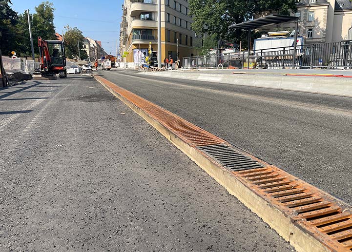 Bauarbeiten auf einer Brücke, im Vordergrund eine Entwässerungsrinnen mit Gussrosten und Bagger, Bauarbeiter, Häuser im Hintergrund