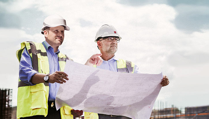 Zwei Bauleiter mit Schutzhelmen und Warnwesten betrachten gemeinsam einen Bauplan auf einer Baustelle.