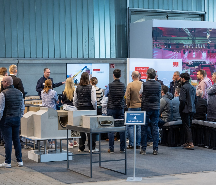Eine Gruppe steht in einer Halle umgeben von Produktdarstellungen und hört einem Redner bei einer Schulung zu.