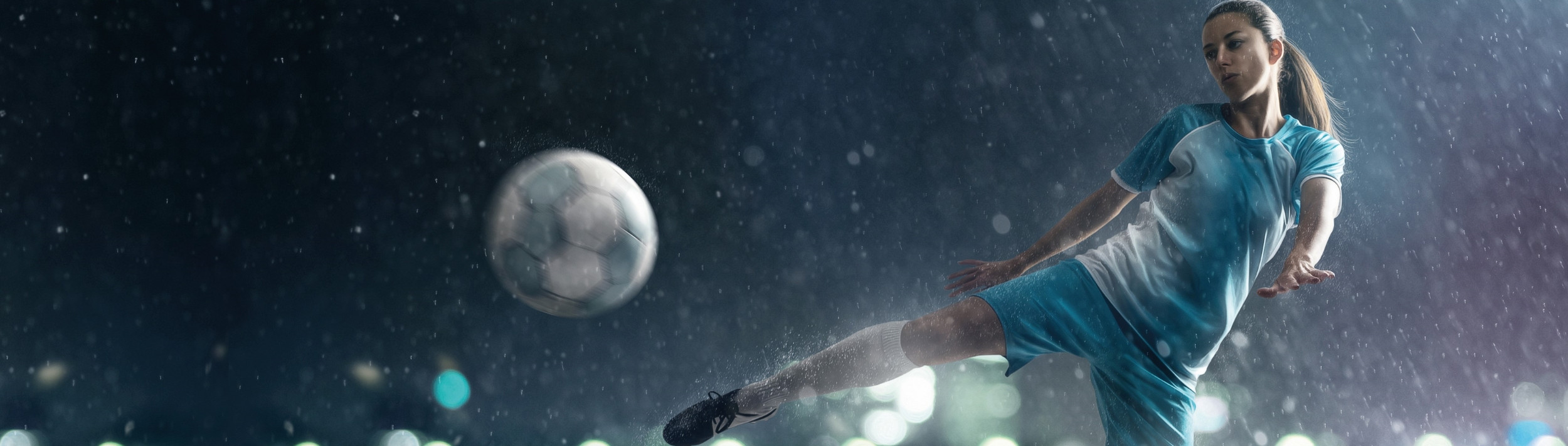 Eine Frau kickt bei Regen und Dunkelheit einen Fußball. Sie ist in Bewegung, ihr Bein ausgestreckt, während sie den Ball trifft.