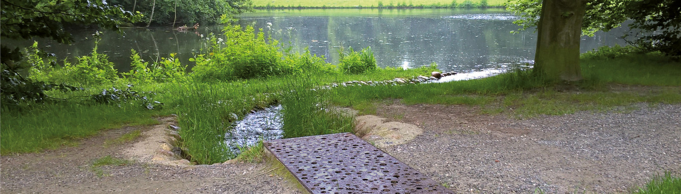 Kleiner Bach fließt zu einem See, umgeben von Bäumen und Gras, im Vordergrund ein Flächenrost.