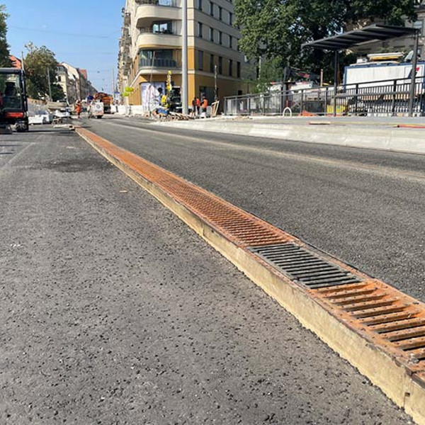 Bauarbeiten auf einer Brücke, im Vordergrund eine Entwässerungsrinnen mit Gussrosten und Bagger, Bauarbeiter, Häuser im Hintergrund