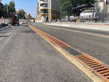 Bauarbeiten auf einer Brücke, im Vordergrund eine Entwässerungsrinnen mit Gussrosten und Bagger, Bauarbeiter, Häuser im Hintergrund