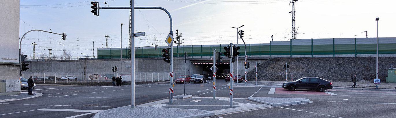 Straßenkreuzung mit Ampeln, mehreren Autos und zwei Fußgängern vor einer Bahnunterführung und Lärmschutzwand.