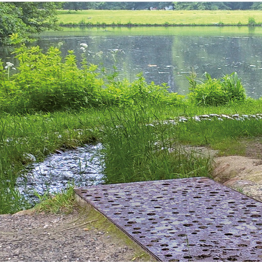 Kleiner Bach fließt zu einem See, umgeben von Bäumen und Gras, im Vordergrund ein Flächenrost.