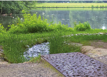 Kleiner Bach fließt zu einem See, umgeben von Bäumen und Gras, im Vordergrund ein Flächenrost.