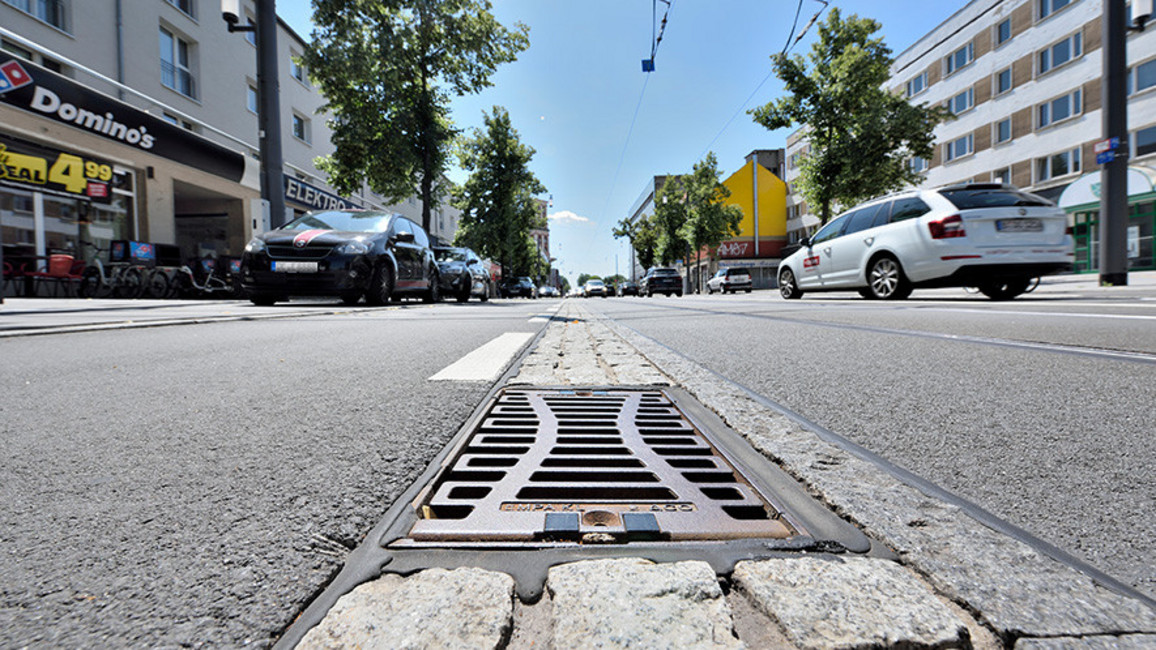 Blick auf eine Straßenentwässerung mit Gitterrost in einer Straße, umgeben von parkenden Autos, Gebäuden und Bäumen bei Sonne.
