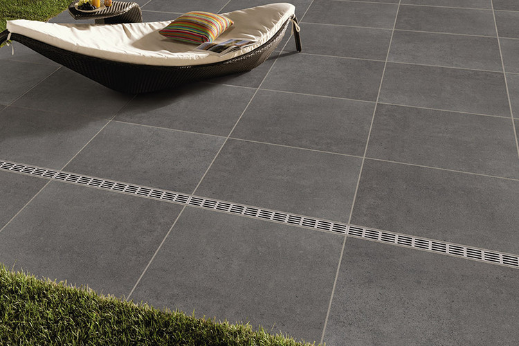 Große Terrasse mit grauen quadratischen Platten, integrierter Entwässerungsrinne und einer modernen Liege mit Kissen.