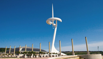 Außenansicht des Olympiastadions in Barcelona, mit seiner klassischen Architektur und eleganten Fassade. Das Stadion hat eine ovale Form und ist von dekorativen Säulen und Bögen umgeben. Die Struktur verbindet historische und moderne Elemente, und im Hintergrund sind die Stadt und grüne Hügel sichtbar.