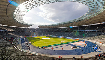 Innenansicht des Olympiastadions in Berlin, mit einer weitläufigen ovalen Spielfläche, umgeben von hohen Tribünen. Die Architektur ist geprägt von klassischen Elementen und einem markanten blauen Laufbahnring. Tageslicht strömt durch das offene Dach.