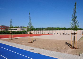 Sportplatzentwässerung ökologisch und ökonomisch wertvoll Blaue Laufbahn vor einem von Bäumen eingefassten Sportplatz. Die Sonne scheint, der Himmel ist blau.