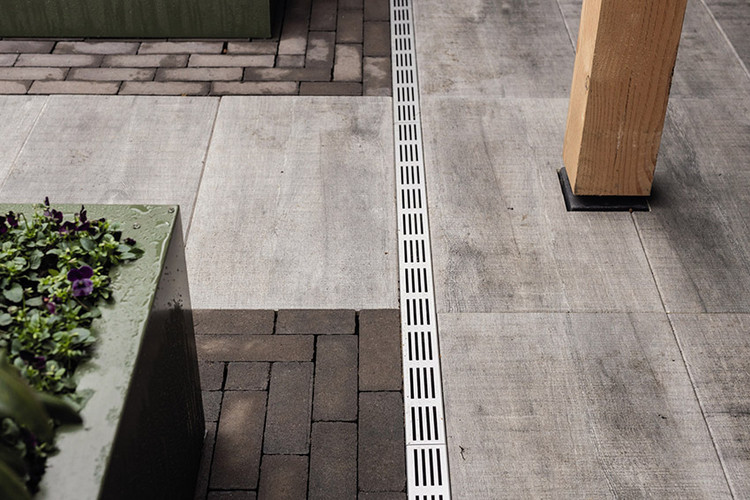 Terrassenbereich mit grauen Platten und dunklen Pflastersteinen, durchzogen von einer schmalen Entwässerungsrinne aus Edelstahl..