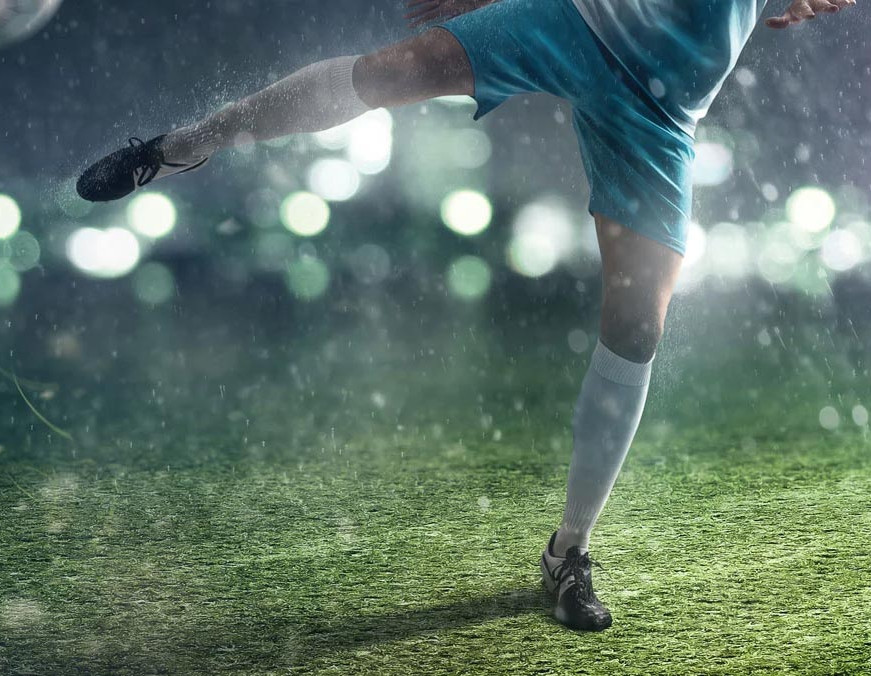 Fußballspieler in blauer Sportkleidung schießt Ball auf nassem Rasen bei Regen, unter Flutlicht in dunkler Umgebung.