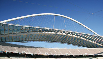 Außenansicht des Olympiastadions in Athen, mit seiner modernen und eleganten Architektur. Das Stadion hat eine ovale Form und ist von einem markanten, geschwungenen Dach aus weißen Stahlträgern bedeckt.
