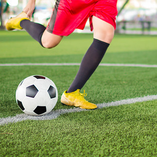 Mann mit roter Sporthose und gelben Fußballschuhen tritt Fußball auf Rasenplatz.