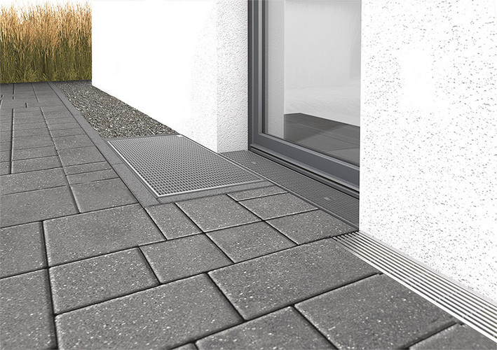 Lichtschacht dezentral vor einer Terrassentür, rechts eine Entwässerungsrinne, links ein schmaler Kiesstreifen, umgeben von Pflastersteinen.