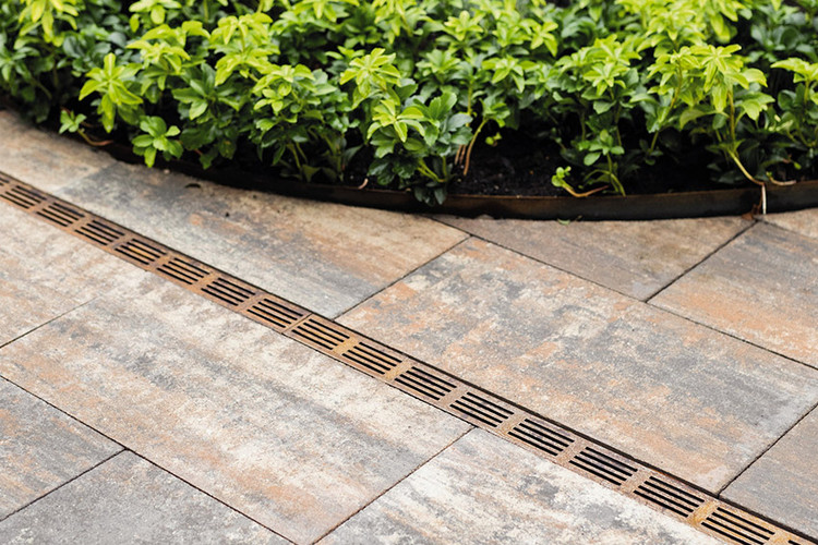 Terrassenplatten in Natursteinoptik mit schmaler Entwässerungsrinne aus Cortenstahl und angrenzendem Beet mit grünen Pflanzen.