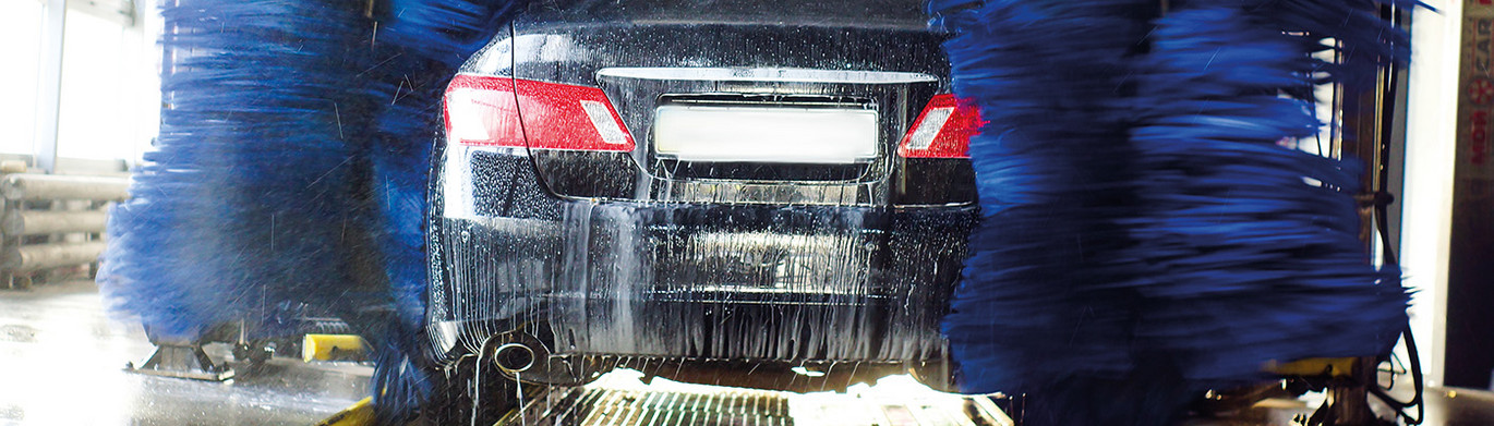 Schwarzes Fahrzeugheck in einer Waschanlage, Wasser und Schaum laufen am Auto herunter, rotierende Bürsten sind sichtbar.