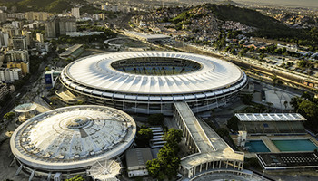 Das Olympiastadion in Rio de Janeiro, ein weitläufiges Sportstadion mit einer ovalen Form, umgeben von Tribünen. Die Architektur ist modern, mit einem offenen Dach und großen Sitzbereichen für Zuschauer. Im Hintergrund sind die Stadt und die umliegenden Hügel zu sehen.